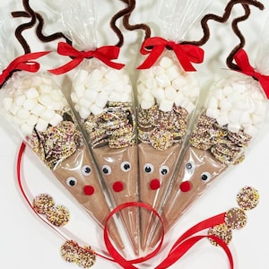 Puede incluir: Cuatro conos de chocolate caliente con temática de reno, envueltos en celofán. Cada cono está lleno de malvaviscos, chocolate y chispas, con ojos saltones y una nariz roja. Cuernos de limpiapipas marrones y cinta roja.
