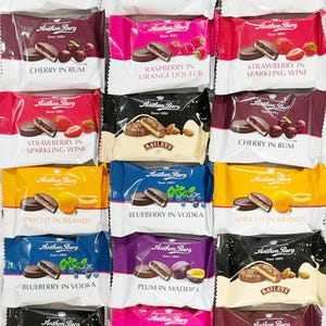 Peut inclure: Assortiment de truffes au chocolat emballées individuellement avec différentes saveurs, notamment cerise au rhum, framboise à la liqueur d'orange, fraise au vin mousseux, abricot à la liqueur de brandy, myrtille à la vodka et prune au madère. Chaque truffe est surmontée d'un disque de chocolat et décorée de fruits et d'autres embellissements. L'emballage porte le nom de la marque "Anthon Berg" et le slogan "Depuis 1884".