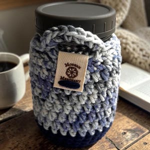 Pode incluir: Um frasco Mason com tampa escura é envolto em uma capa de crochê feita à mão. A capa apresenta uma mistura de fios azuis, cinza e branco, com uma base azul escura. Uma pequena etiqueta com o texto "Modern Merchant" está anexada.