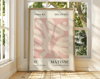 Op de afbeelding: Een ingelijste kunstdruk met een Matisse-ontwerp. Het kunstwerk toont abstracte vormen in lichtroze en gebroken wit tegen een roze achtergrond. De tekst bovenaan luidt "PAPIERS DÉCOUPÉS" en onderaan "MATISSE PARIS, FRANCE 1955".