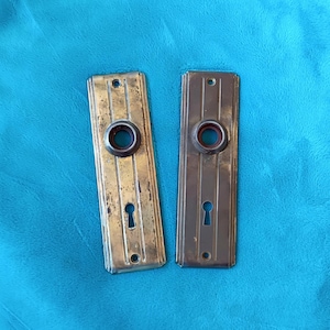 May include: Two rectangular metal door plates with a vertical striped design. Each plate features a central circular opening for a doorknob and a smaller keyhole opening below it, with screw holes at the top and bottom of each plate.