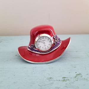 May include: A red hat-shaped clock with a silver-tone clock face. The clock face has the brand name "Elgin" printed on it. The hat has a purple band and floral accents. The clock is a decorative item.
