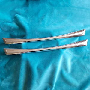 May include: Two polished, silver-toned cabinet handles with a curved design and angled ends. The handles are set against a textured, teal-colored fabric background, creating a contrast in color and texture. These handles are likely for furniture.