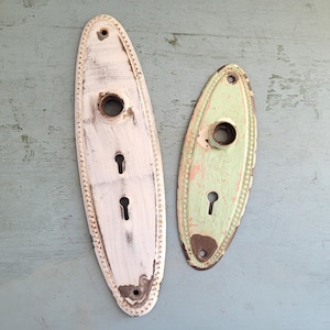 May include: Two vintage door plates. One is a long, oval shape painted white with a dotted border and two keyholes. The other is a smaller, oval shape painted light green with a keyhole. Both have distressed paint.