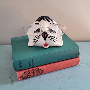 May include: A ceramic dog-shaped eyeglass holder with clear-framed glasses, resting on a stack of two books. The dog has black and white markings, a red mouth, and black spots. The books are teal and red.