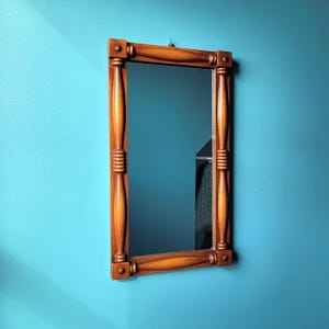 May include: A rectangular wooden mirror with a light brown frame. The frame has decorative vertical columns and horizontal bars at the top and bottom. The mirror is hanging on a teal wall.