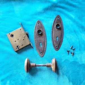 May include: Antique brass door hardware set, including a mortise lock, two oval backplates with keyholes, a pair of doorknobs, and several screws. The hardware is arranged on a turquoise background.