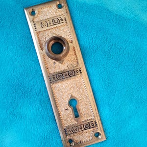 May include: A vintage brass door plate with a keyhole and decorative detailing. The rectangular plate has a textured surface and is adorned with intricate patterns and designs. It features a circular opening and four screw holes.