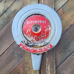 May include: A vintage Oldsmobile Sky Rocket air cleaner with a red and silver finish. The air cleaner has the words "OLDSMOBILE" and "Sky Rocket" in white lettering. The words "ULTRA HIGH COMPRESSION" are also visible.