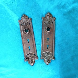 May include: Two rectangular bronze door plates featuring ornate, raised scrollwork patterns at the top and bottom. Each plate includes a circular opening for a door knob and two vertical keyhole slots, showcasing a weathered, aged metal finish.