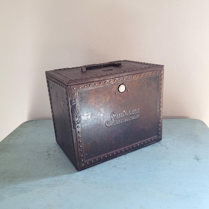May include: Vintage Sunbeam Electric Iron box, a rectangular metal container with a handle on top. The box is a dark, aged brown with visible rivets and the words "Sunbeam Electric Iron" embossed on the front. It sits on a light blue surface.