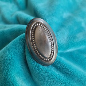 May include: An oval-shaped metal ring featuring a smooth, raised center panel surrounded by a decorative border of small, uniform beads. The metal has a muted, matte grey finish with subtle dark shading along the edges of the oval frame.