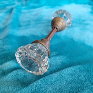 May include: A vintage door knob with clear, faceted glass knobs and a brass-toned metal stem and base. The door knob is set against a teal blue fabric background.