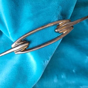 May include: A gold-toned metal hair clip with an abstract design. The clip has a central oval shape with a pointed bar extending from each end. The clip is set against a teal blue, plush background.