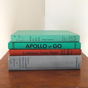 May include: A stack of four hardcover books. The top book is light green with the text West of the Sun by Edgar Pangborn. The second book is bright green with the text Apollo at Go. The third is orange with Earthman, Come Home. The bottom is gray.