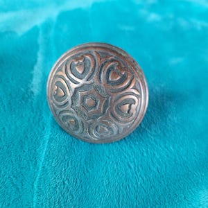 May include: A round, antique-style copper button with an ornate design. The button features a central star shape surrounded by heart-shaped patterns. The button is set against a teal background.