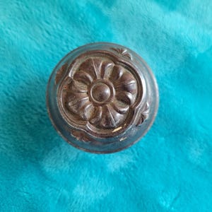 May include: A detailed, antique-style metal cabinet knob with a floral design. The knob is circular with a raised, ornate pattern in shades of brown and silver. The background is a vibrant teal color.
