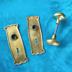 May include: A set of brass door hardware consisting of two rectangular backplates with scalloped edges, each featuring a central circular opening and a keyhole, alongside a single brass door knob with a connecting metal spindle shaft.