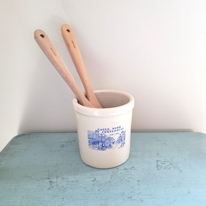 May include: A cream-colored ceramic crock with a blue graphic and text that reads "EAGLE, NEBR CENTENNIAL 1886-1986" holds two wooden spoons. The spoons are light brown and have a hole in the handle. The crock sits on a light blue surface.