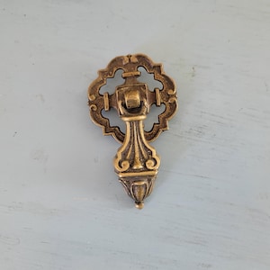 May include: A brass drawer pull with an ornate design. The pull has a decorative, pointed bottom and a round top with a keyhole.