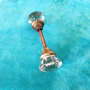 May include: A pair of clear, faceted glass door knobs connected by a central metal spindle. Each knob features a circular, multi-faceted design with a brass-toned metal base that shows signs of aged patina and oxidation on the connecting rod.