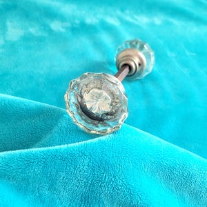 May include: A pair of clear, faceted glass doorknobs with a silver-toned metal base and a dark brown connecting rod. The doorknobs are set against a vibrant turquoise fabric background.