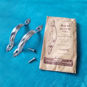 May include: Two polished chrome drawer pulls with screws and a vintage Amerock Drawer Pull package. The package is tan with black text and illustrations. The drawer pulls are curved and shiny, reflecting the light. The package reads "American Cabinet Hardware".