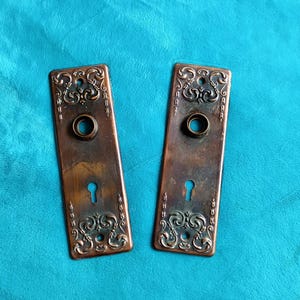 May include: Two rectangular metal door plates with a weathered copper finish. Each plate features an ornate embossed scrollwork pattern at the top and bottom, a central circular opening for a door knob, and a keyhole opening near the bottom.