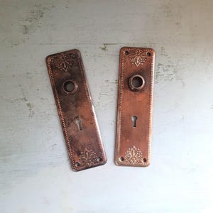 May include: Two antique copper-toned door plates. Each rectangular plate features a keyhole, a circular knob opening, and decorative floral accents. The plates have a vintage, aged appearance, with a slightly tarnished finish.