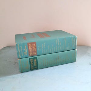 May include: Two vintage turquoise "Modern Encyclopedia of Cooking" books stacked on top of each other. The books have gold lettering and red accents. The books are on a light blue surface.