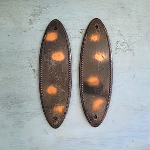 May include: Two antique, oval-shaped door plates with a dark bronze finish. Each plate has a series of small, raised dots around the edge and three lighter, orange-toned spots. The plates are slightly distressed, suggesting age and use.
