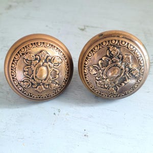 May include: Two antique bronze door knobs. Each knob has a circular design with an ornate floral and oval center pattern. The knobs are a warm, aged bronze color, suggesting vintage or retro style.