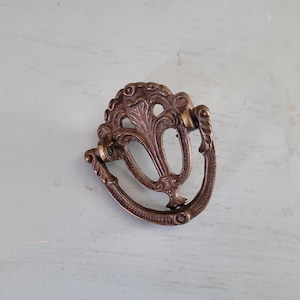 May include: Ornate bronze-toned drawer pull with a decorative floral design. The pull features a curved handle and a detailed backplate with a central leaf motif. A vintage-style hardware piece.