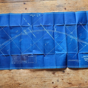 May include: A folded blue blueprint with white lines and text, likely a map or technical drawing. The blueprint is on a wooden surface. The text includes the name of a company and other technical details.