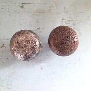 May include: Two antique bronze cabinet knobs with intricate embossed patterns. Each knob is round with a central square design and ornate detailing. The knobs are displayed on a distressed white surface.