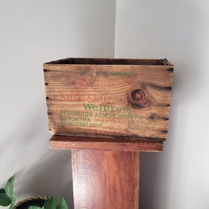 May include: A weathered wooden crate with visible knots and grain. The crate has faded green text that reads "Wetproof" and "Remington Arms Company." The crate is sitting on a wooden stand.