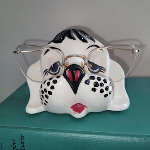 May include: A ceramic dog-shaped glasses holder, painted white with black spots and accents. The dog has blue eyes, a red nose and mouth, and is wearing a pair of silver-rimmed glasses. The holder sits on a green book.