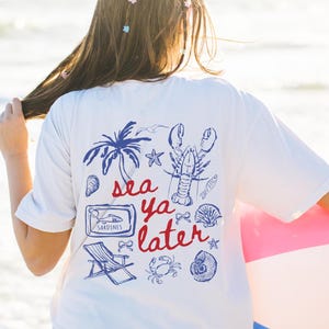 May include: A white t-shirt with a blue and red graphic print that says 'sea ya later'. The graphic includes a lobster, a palm tree, a beach chair, a seashell, a crab, and a can of sardines.