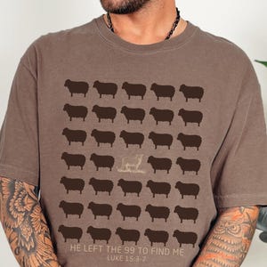 May include: Espresso-colored t-shirt featuring a pattern of brown sheep, with one sheep in a lighter shade. The text "HE LEFT THE 99 TO FIND ME LUKE 15:3-7" is printed below the sheep design. The shirt is a casual wear item.