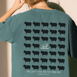 May include: A blue-green t-shirt featuring a grid of black sheep silhouettes on the back. One white sheep outline is centered among them. Below the grid, the text reads: HE LEFT THE 99 TO FIND ME LUKE 15:3-7 in white capital letters.