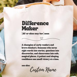 May include: Cream-colored tote bag with the words "Difference Maker" in a bold, black font. The definition of "Difference Maker" is printed below. The bag has long handles and is held by a person sitting on the grass.