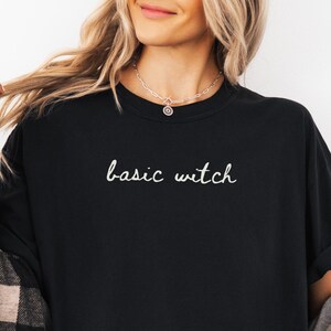 May include: A black crew neck t-shirt featuring the white text basic witch printed in a cursive font on the chest. The shirt is worn under an unbuttoned black and tan plaid flannel shirt with long sleeves and a collar.