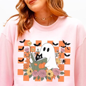 May include: Light pink sweatshirt featuring a Halloween-themed graphic. The graphic includes a white ghost reading a book with a black cat, surrounded by flowers, bats, and a spider, set against an orange and white checkered background.