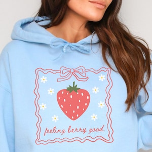 May include: A light blue hoodie with a graphic of a strawberry in a pink frame with white flowers and the text "feeling berry good".