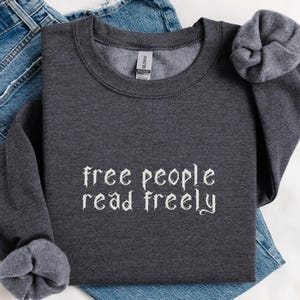 May include: Dark heather gray sweatshirt with the words 'free people read freely' in white, stylized text. The sweatshirt is folded over a pair of blue jeans. The sleeves are cuffed. A dark heather circle with the words 'DARK HEATHER' is in the bottom right corner.