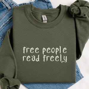 Może przedstawiać: Oliwkowozielona bluza z napisem "free people read freely" gotycką czcionką. Bluza jest złożona na parze niebieskich dżins&oacute;w. Kolor bluzy oznaczony jest jako "Military Green".