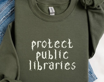 May include: An olive green sweatshirt with the text "protect public libraries" in a gothic font. The sweatshirt is folded on top of a pair of blue jeans. The sleeve is knotted. The color is labeled "military green".
