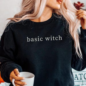 May include: Black sweatshirt with the text "basic witch" in white. The sweatshirt is worn with blue jeans. A white mug is held in the hand. A pumpkin pillow is in the background.