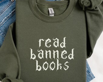 May include: Military green sweatshirt with the text "read banned books" in a gothic font. The sweatshirt is folded on top of a pair of blue jeans. The sleeve is knotted at the end.