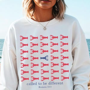 May include: White sweatshirt with a pattern of red lobsters, one blue lobster, and the text "called to be different" from Romans 12:2. The sweatshirt is a casual style.